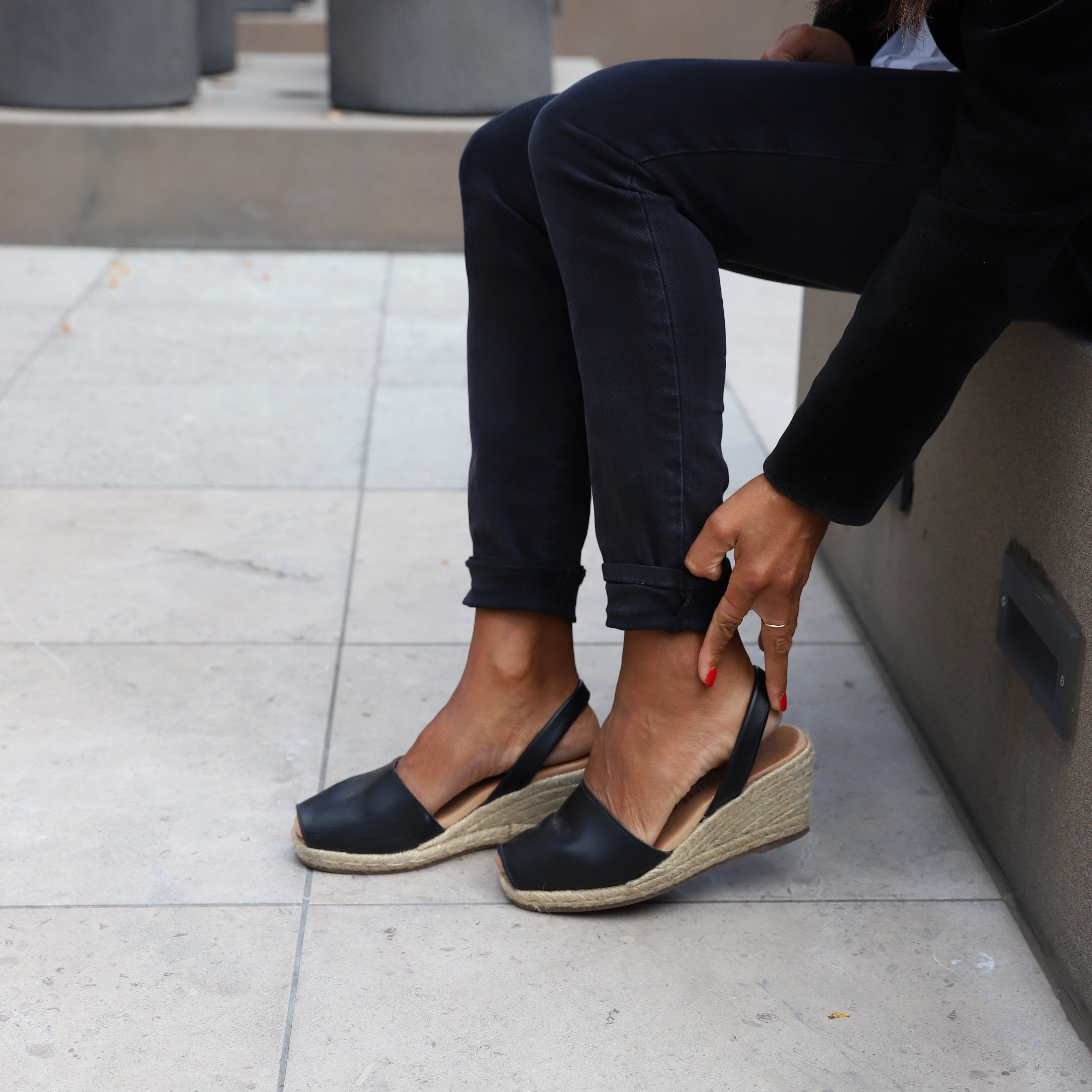 woman sitting down wearing black pants and putting on wedge espadrille sandals in black